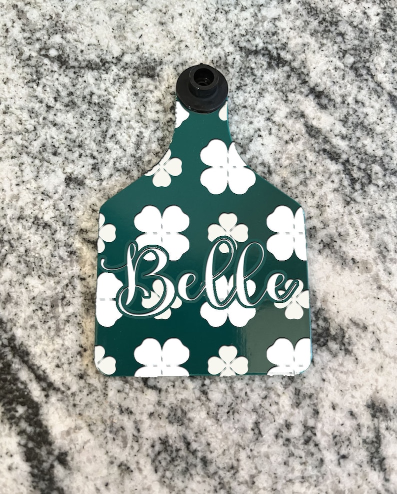 May include: Green plastic livestock ear tag with a white four-leaf clover pattern and the name "Belle" in white cursive lettering.