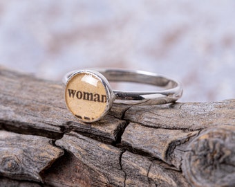 May include: A silver ring with a round, beige resin cabochon featuring the word "woman" in black lettering.