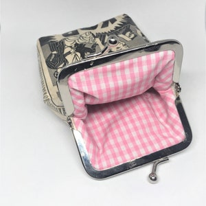 May include: A small, open coin purse with a pink and white gingham lining. The exterior of the purse is made of a cream-colored fabric with a black and white comic book illustration.