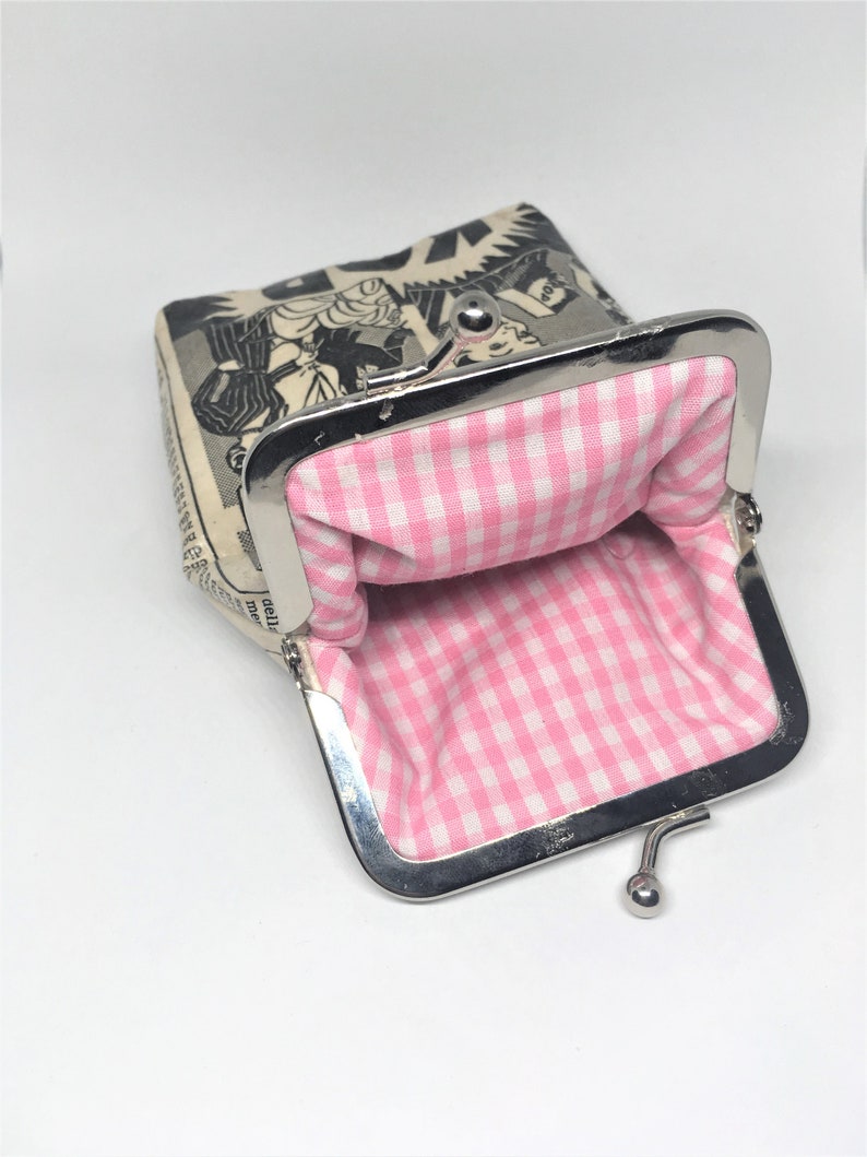 May include: A small, open coin purse with a pink and white gingham lining. The exterior of the purse is made of a cream-colored fabric with a black and white comic book illustration.