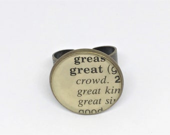 Puede incluir: Un anillo de bronce con una parte superior redonda de vidrio crema. La parte superior de vidrio presenta texto negro de una página de diccionario, que dice "great (g... crowd. 2 great kin great si... good"