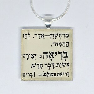 May include: Square pendant with Hebrew text, suspended from a silver chain. The pendant has a cream-colored background with black lettering. The text appears to be religious or spiritual in nature.