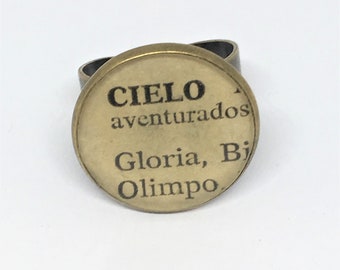 Puede incluir: Un anillo dorado con una parte superior redonda de vidrio. La parte superior presenta texto negro que dice "CIELO aventurados Gloria, Bi Olimo."