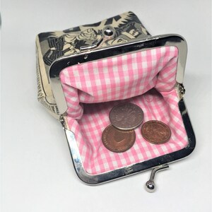 May include: A small, black and white comic book print coin purse with a pink and white gingham lining. The purse is open and has three coins inside.