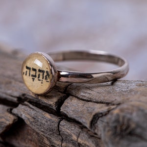 Pode incluir: Um anel de prata com uma pedra redonda de cor creme com texto em hebraico que diz "Mah Tov".