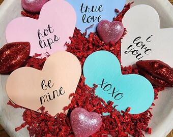 May include: A bowl filled with red shredded paper and six painted wooden hearts. The hearts have different colors and phrases written on them, including "true love", "hot lips", "be mine", "xoxo", "i love you", and "hot lips".