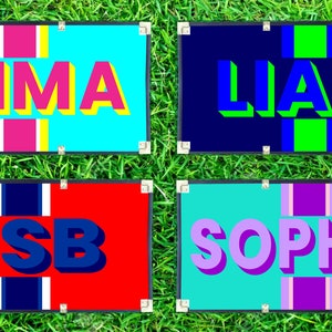 May include: Four rectangular signs with names and initials. "EMMA" in pink and yellow on a turquoise background. "LIAM" in blue and green on a navy background. "CSB" in blue on a red background. "SOPHIA" in purple on a teal background.