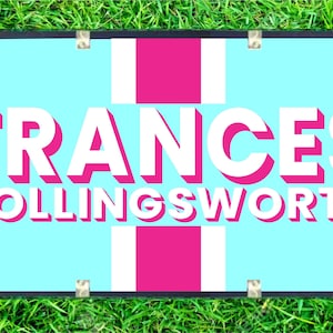 May include: A rectangular sign with the name "FRANCES HOLLINGSWORTH" in white, outlined in pink, on a light blue background. The sign has a pink cross design and is framed in black with silver corner protectors. The sign is placed on a bed of green grass.