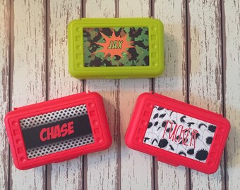 May include: Three personalized pencil cases. One is green with a camouflage pattern and the name "JAX" in a red speech bubble. The other two are red. One has a black and white pattern with the name "CHASE" in white. The other has a white and black shark pattern with the name "TUCKER" in white.
