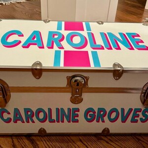 May include: A white trunk with the name "CAROLINE" in turquoise letters with a pink shadow and a pink stripe. The name "CAROLINE GROVES" is also displayed on the trunk in the same color scheme. The trunk has metal hardware.