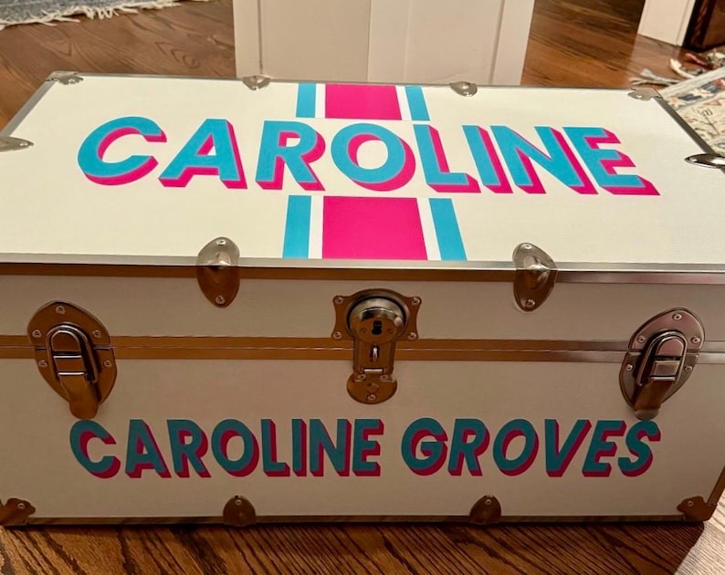 May include: A white trunk with the name "CAROLINE" in turquoise letters with a pink shadow and a pink stripe. The name "CAROLINE GROVES" is also displayed on the trunk in the same color scheme. The trunk has metal hardware.