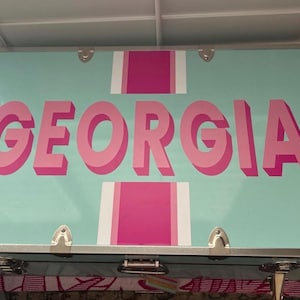 May include: A light blue trunk with the name "GEORGIA" in large, pink, 3D letters. The trunk has silver hardware and pink and white stripes. The trunk is likely used for storage or travel.