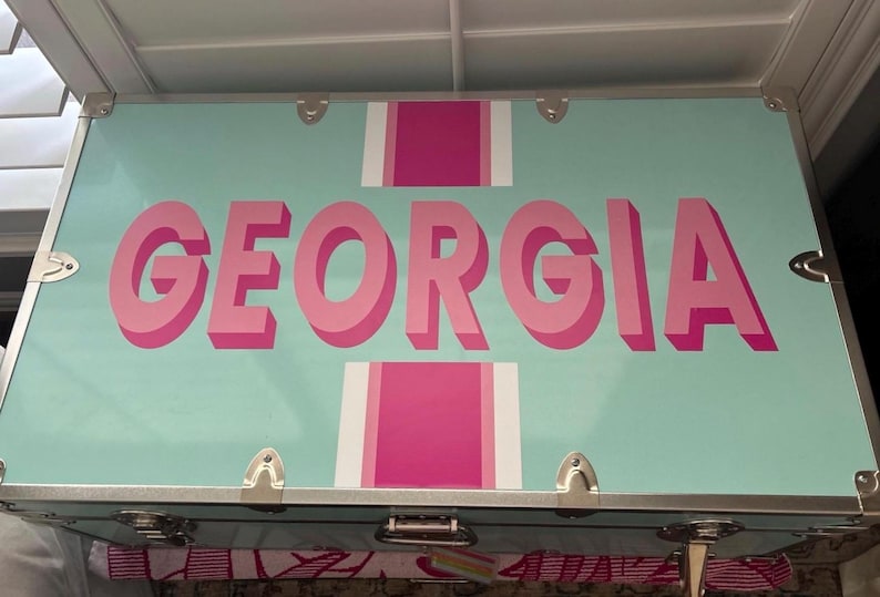 May include: A light blue trunk with the name "GEORGIA" in large, pink, 3D letters. The trunk has silver hardware and pink and white stripes. The trunk is likely used for storage or travel.