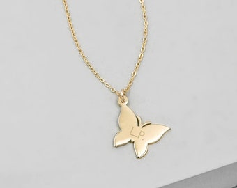 May include: A gold-colored butterfly pendant necklace. The butterfly pendant has the initials "L.P." engraved on it. The necklace chain is delicate and gold-colored. The background is a light gray.