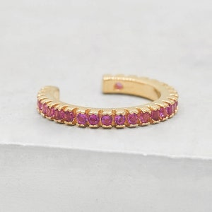 May include: A gold-colored ear cuff with a row of small, bright pink gemstones. The cuff is open at one end and sits on a white surface. The gemstones are evenly spaced along the front of the cuff.