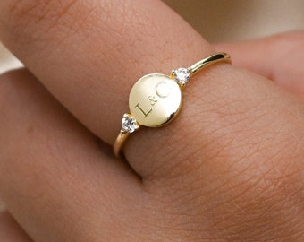 May include: A gold ring featuring a small circular disc engraved with the letters L&C. The disc is flanked on either side by a single clear, round-cut gemstone set into the thin gold band, creating a delicate and polished appearance.