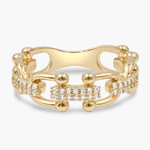 May include: A gold-colored ring with a unique design. The ring features a series of connected segments, each adorned with small, sparkling stones. The overall shape is a continuous loop, with a central decorative element. The ring is set against a plain white background.