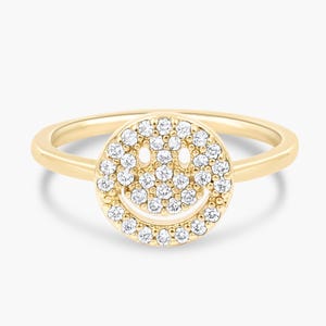 May include: A gold-colored ring featuring a smiley face design. The face is made of small, sparkling white gemstones. The ring band is a thin, polished gold. The overall design is simple and elegant, suitable for everyday wear.