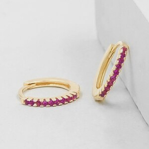 May include: A pair of gold hoop earrings featuring a row of small, vibrant pink gemstones set along the front edge. One earring lies flat while the other stands upright, showcasing the hinged closure mechanism and the polished gold metal finish.