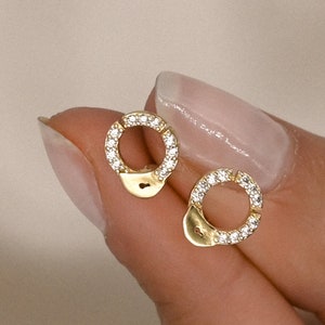 Puede incluir: Un par de pendientes de botón dorados con forma de esposas. Cada pendiente presenta un círculo incrustado con pequeños cristales transparentes. Los pendientes están sostenidos por los dedos de una persona sobre un fondo neutro.