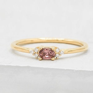 May include: A delicate gold ring featuring a pink oval gemstone at the center, accented by small, clear gemstones. The ring has a thin band and a minimalist design, perfect for everyday wear.