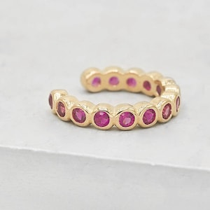 May include: A gold-colored ear cuff featuring a series of vibrant pink gemstones. The cuff curves to fit the ear, with each gemstone set in a bezel setting. The design is simple yet elegant, suitable for everyday wear or special occasions.
