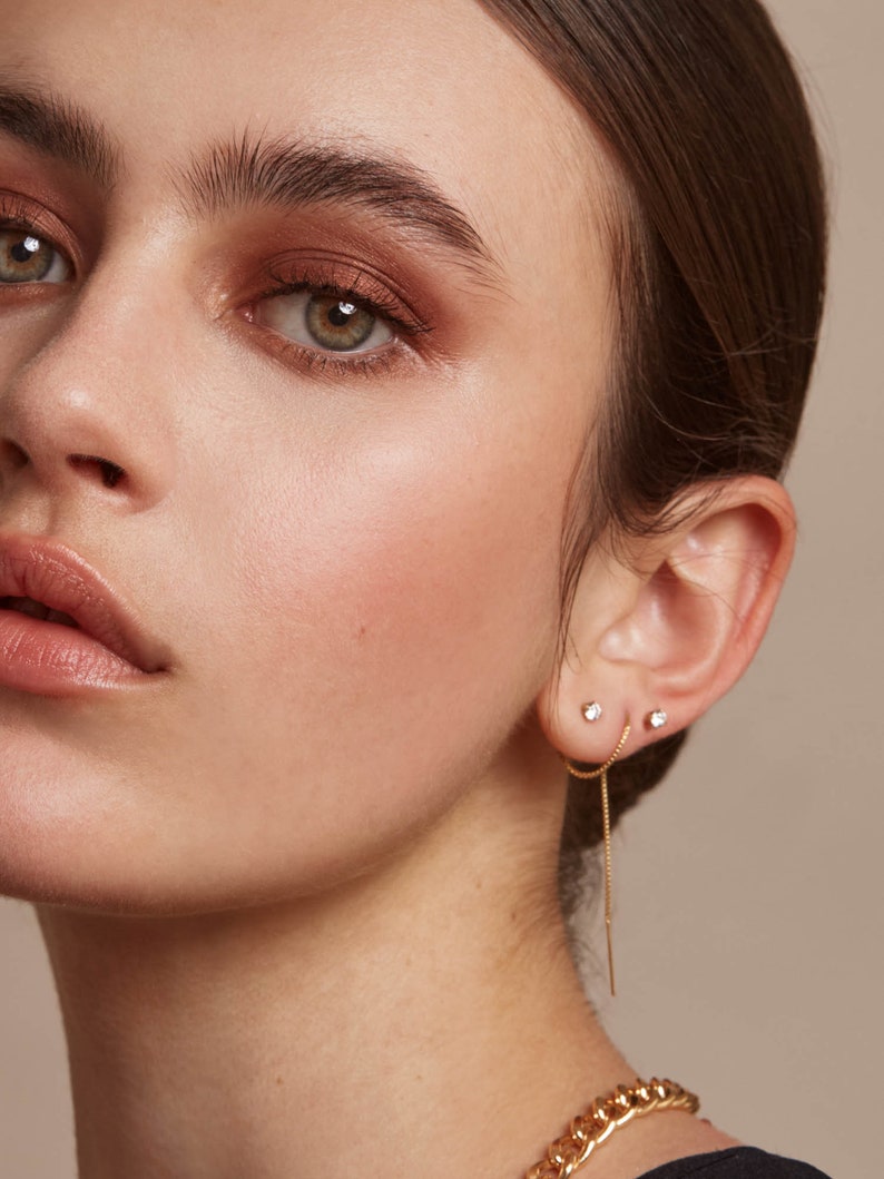May include: Close-up of a person's face, showcasing three small, sparkling stud earrings. One is a classic stud, and the other is a chain-style earring. A gold chain necklace is visible at the bottom of the frame. The person has light skin and is wearing makeup.