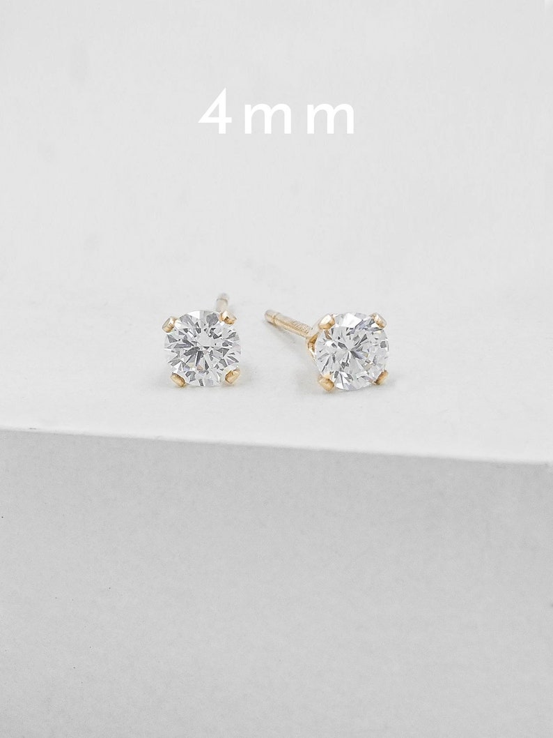 May include: A pair of gold stud earrings, each featuring a clear, round, 4 mm gemstone. The earrings are set in a simple, classic design, perfect for everyday wear. The background is a clean, white surface.
