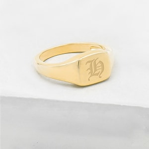 May include: A gold-colored signet ring with a square face. The ring features an engraved letter "H" in an old English font. The ring is set against a white background.