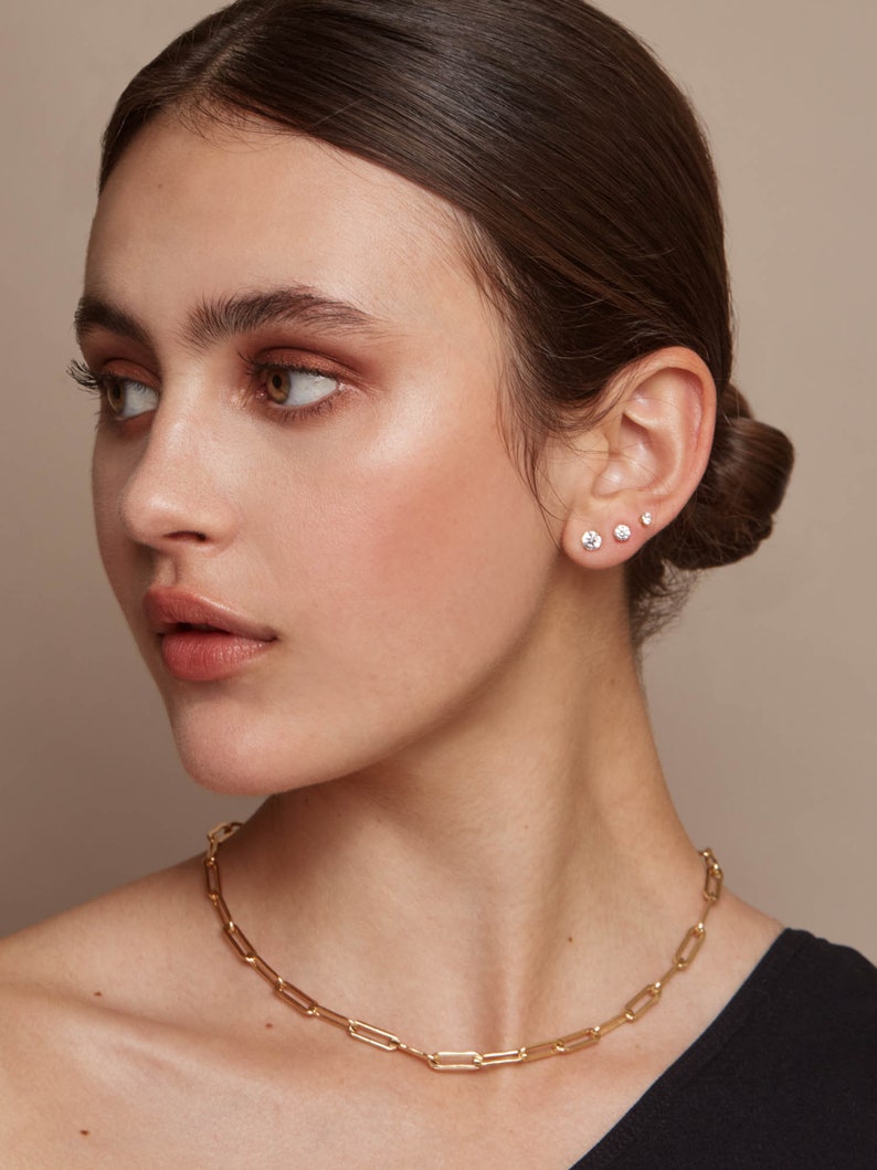 May include: A gold-colored paperclip chain necklace and three small stud earrings. The earrings are set in the ear. The necklace is a simple, elegant design. The model is wearing a black top.