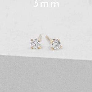 May include: A pair of gold stud earrings with a clear, round, 3 mm gemstone. The earrings are set on a white surface, with the text "3 mm" above them.
