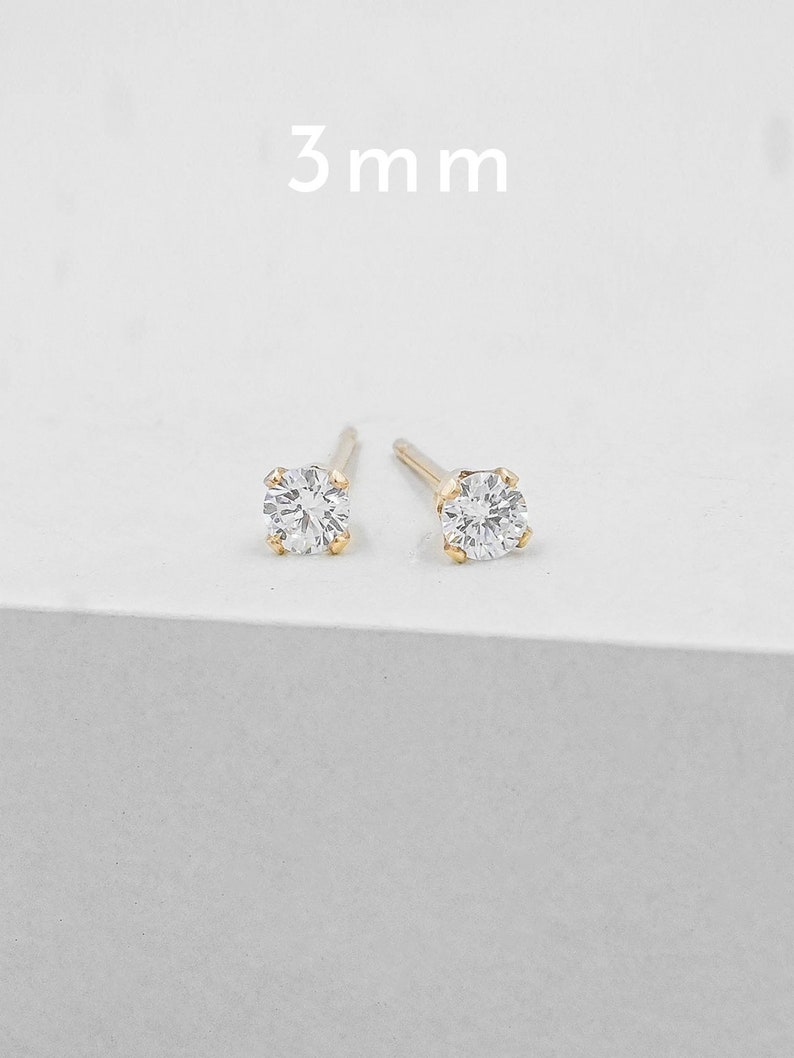 May include: A pair of gold stud earrings with a clear, round, 3 mm gemstone. The earrings are set on a white surface, with the text "3 mm" above them.