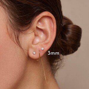 May include: A close-up of an ear with two small, round, diamond stud earrings and a gold chain earring. The diamond studs are 3mm in size. The chain earring dangles down from the ear.