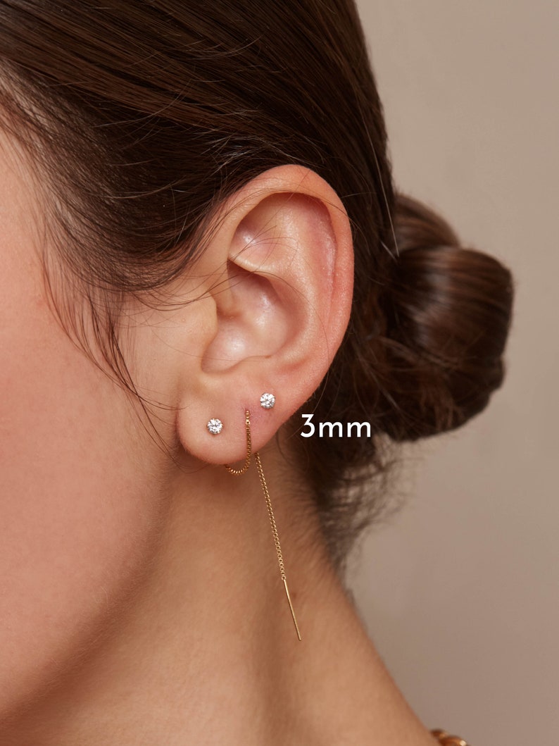 May include: A close-up of an ear with two small, round, diamond stud earrings and a gold chain earring. The diamond studs are 3mm in size. The chain earring dangles down from the ear.