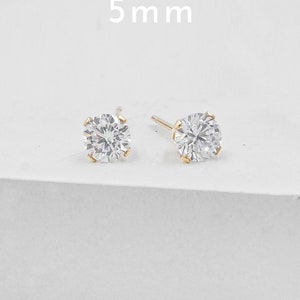 May include: Two gold stud earrings with a clear, round, faceted stone. The earrings are 5 mm in size. The studs are set in a gold setting and are displayed on a white background.