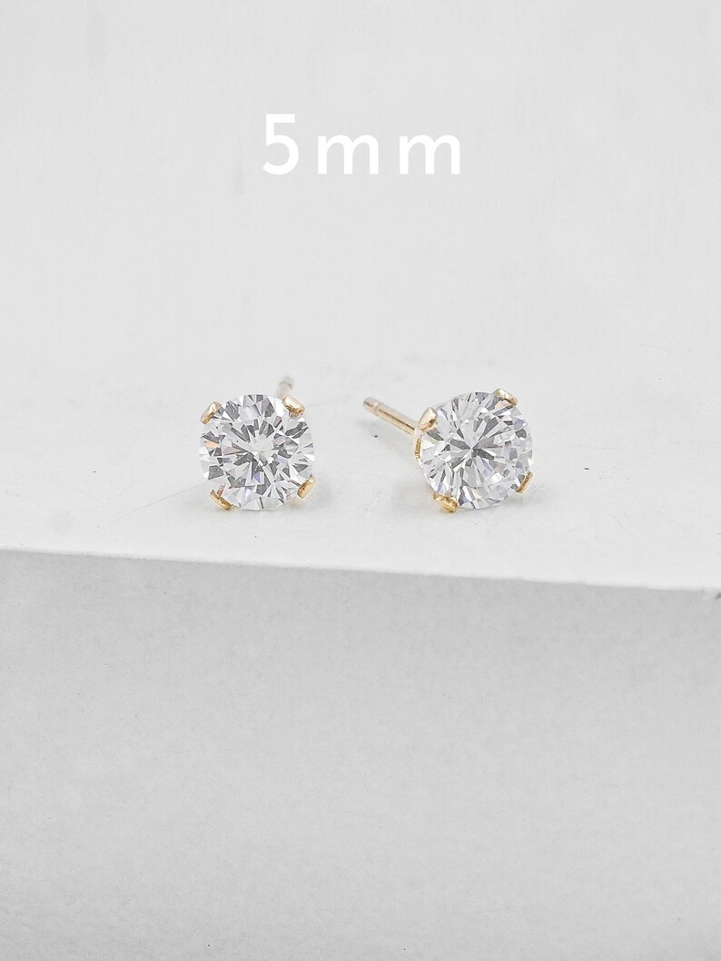 May include: Two gold stud earrings with a clear, round, faceted stone. The earrings are 5 mm in size. The studs are set in a gold setting and are displayed on a white background.