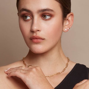 May include: A woman wearing gold jewelry, including a necklace, bracelet, and ring. The necklace and bracelet feature a chain link design. The woman also has two sets of small stud earrings. The jewelry is set against a neutral background.