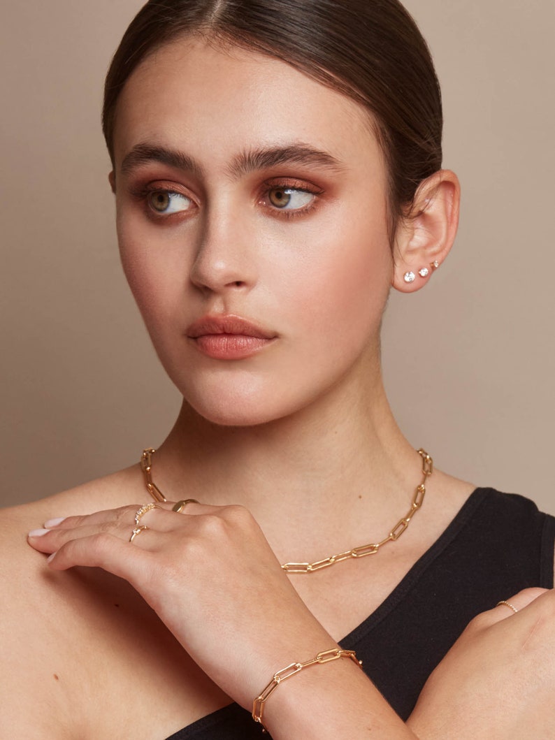 May include: A woman wearing gold jewelry, including a necklace, bracelet, and ring. The necklace and bracelet feature a chain link design. The woman also has two sets of small stud earrings. The jewelry is set against a neutral background.