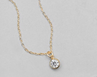 May include: A delicate gold-colored necklace with a small, round, clear gemstone pendant. The pendant is set in a gold bezel and hangs from a fine chain. The necklace is displayed against a light gray background.