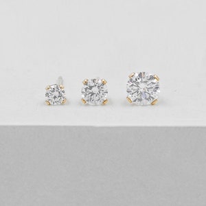 May include: Three clear, round-cut diamond stud earrings of varying sizes. The diamonds are set in gold-colored prongs. The earrings are displayed on a white surface, showcasing their sparkle and clarity.