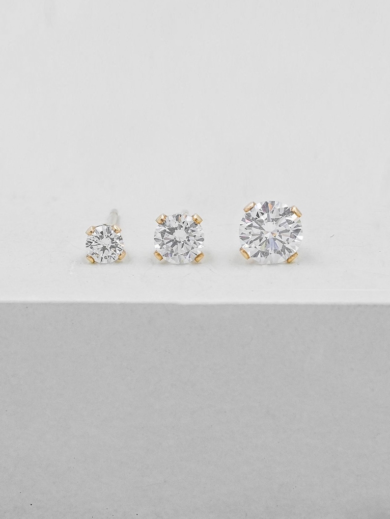 May include: Three clear, round-cut diamond stud earrings of varying sizes. The diamonds are set in gold-colored prongs. The earrings are displayed on a white surface, showcasing their sparkle and clarity.