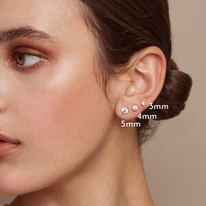 May include: A close-up of a person's ear adorned with three clear, round stud earrings. The earrings are 3mm, 4mm, and 5mm in diameter. The skin tone is fair, and the background is a soft, neutral color.