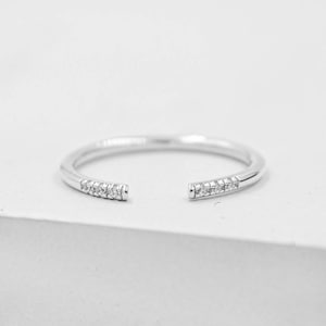 May include: A silver-colored open ring with small, clear gemstones set on each end. The ring has a minimalist design and is set against a white background. The ring is a piece of jewelry.