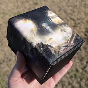 Könnte beinhalten: Eine schwarze Deck-Box mit einem kontrastreichen, detaillierten Grafikdruck auf dem Deckel. Das Bild zeigt eine dunkle, felsige Landschaft mit einem zentralen leuchtenden Wasserfall, hellen Lichteffekten und texturiertem Gelände in Schwarz, Weiß und Gold.