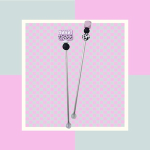 May include: Two silver stirring sticks with decorative tops. One top has a pink and white cow print design with the words "Howdy Y'all" in white. The other top is a pink and white cube.