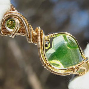 May include: A handcrafted pendant featuring a vibrant green gemstone encased in intricate gold-colored wire. The design includes a smaller green stone and delicate wire wrapping, creating a unique and artistic jewelry piece.