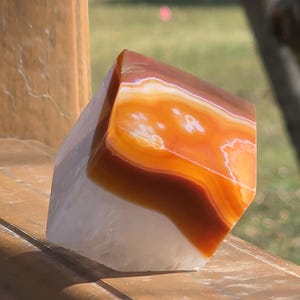 May include: A polished, geometric gemstone with layers of orange, brown, and white. The stone is displayed on a wooden surface, with a blurred green background. The stone's facets catch the light, showcasing its natural patterns.