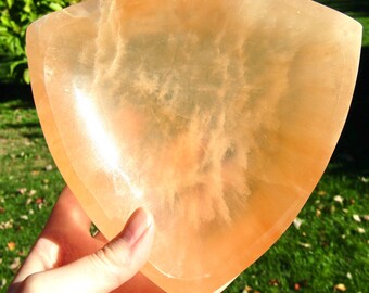 May include: A translucent, orange-hued selenite bowl with a rounded, shield-like shape. The bowl is held in a hand, with the background showing green grass and foliage. The bowl's surface has a subtle, cloudy pattern.