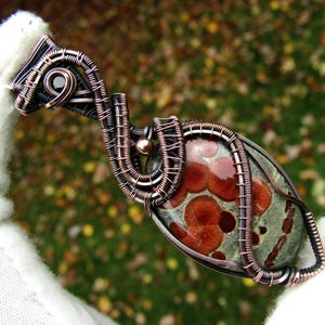 Op de afbeelding: Een met draad omwikkelde hanger met een ovale orbiculaire jaspissteen met roodbruine cirkelvormige patronen op een saliegroene achtergrond. De steen is gevat in donker, geoxideerd koperdraad met ingewikkelde wikkelingen en vlechtwerk.