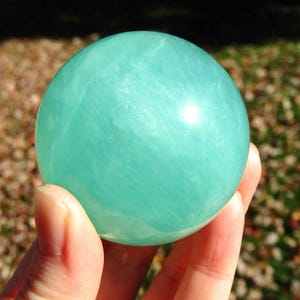 May include: A smooth, round, light teal-colored sphere. The sphere is held in a hand, with the background out of focus. The sphere has a glossy finish and appears to be made of a translucent material. The number "2" is in the upper right corner.